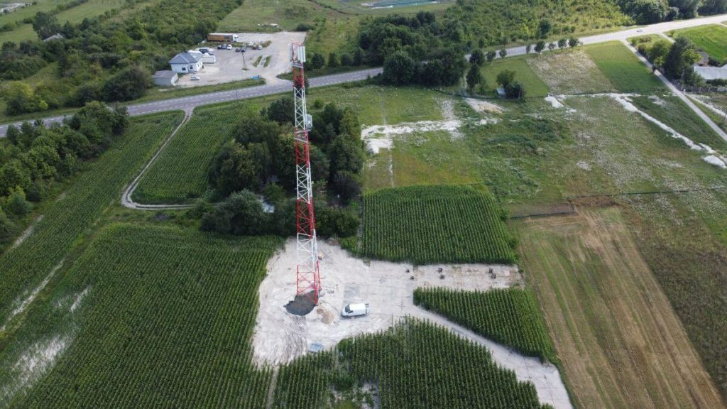 Wieża telekomunikacyjna na tle pól uprawnych i zadrzewień, z pobliskimi budynkami i drogą.