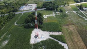 Wieża telekomunikacyjna na tle pól uprawnych i zadrzewień, z pobliskimi budynkami i drogą.