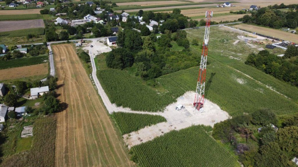 Wiejska sceneria z zielonymi polami, mnóstwem domów, a w centralnej części wieża telekomunikacyjna.