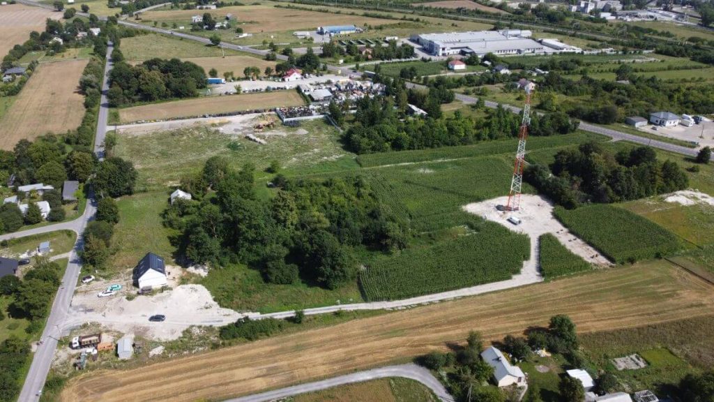 Widok z lotu ptaka na teren wiejski z drzewami, polem i małymi budynkami oraz wieżą telekomunikacyjną.