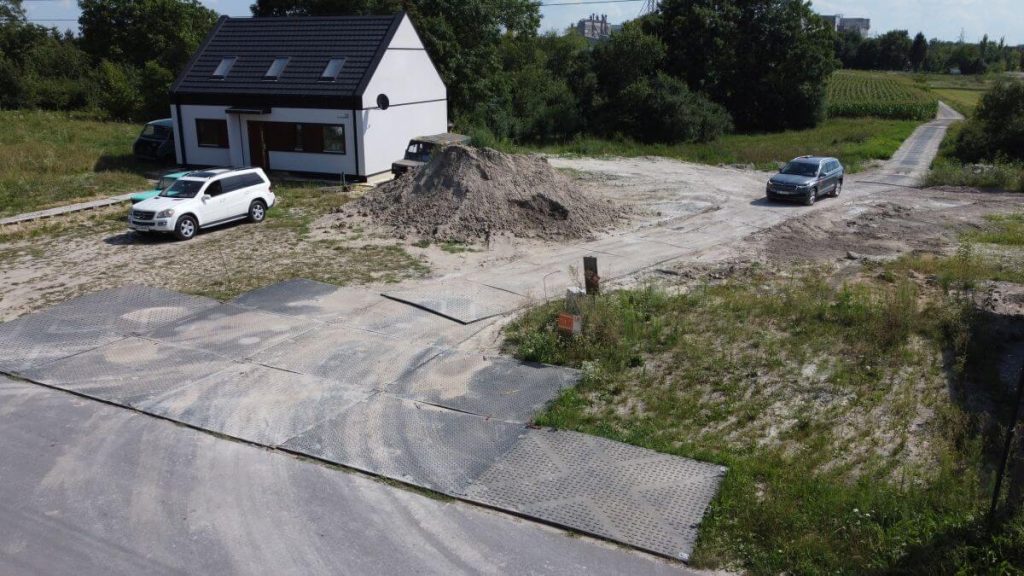 Na zdjęciu widać nowoczesny dom z białą elewacją, samochody, a także plac budowy z hałdą piachu. Otoczenie to tereny zielone.